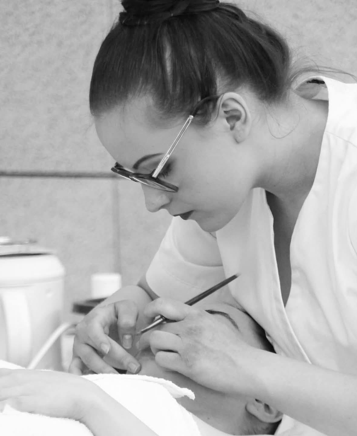 Clémence, en blouse blanche, réalise un soin du regard au pinceau sur une cliente allongée